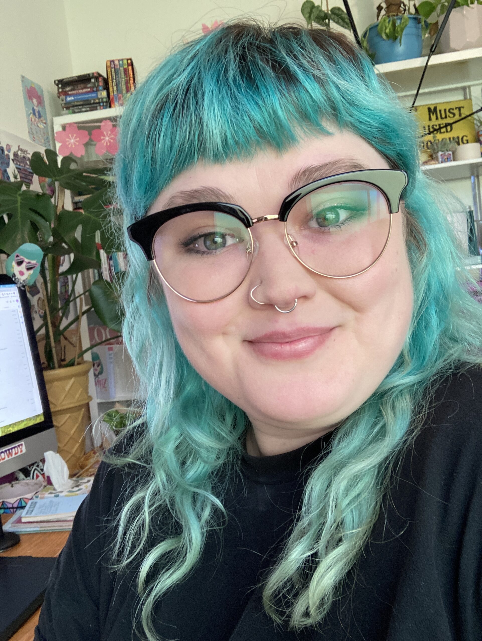 Photograph of Emma 'Bambz' Beukers. Emma is smiling, has blue tinted hair and is wearing glasses with black rims at the top. Emma is in her studio.